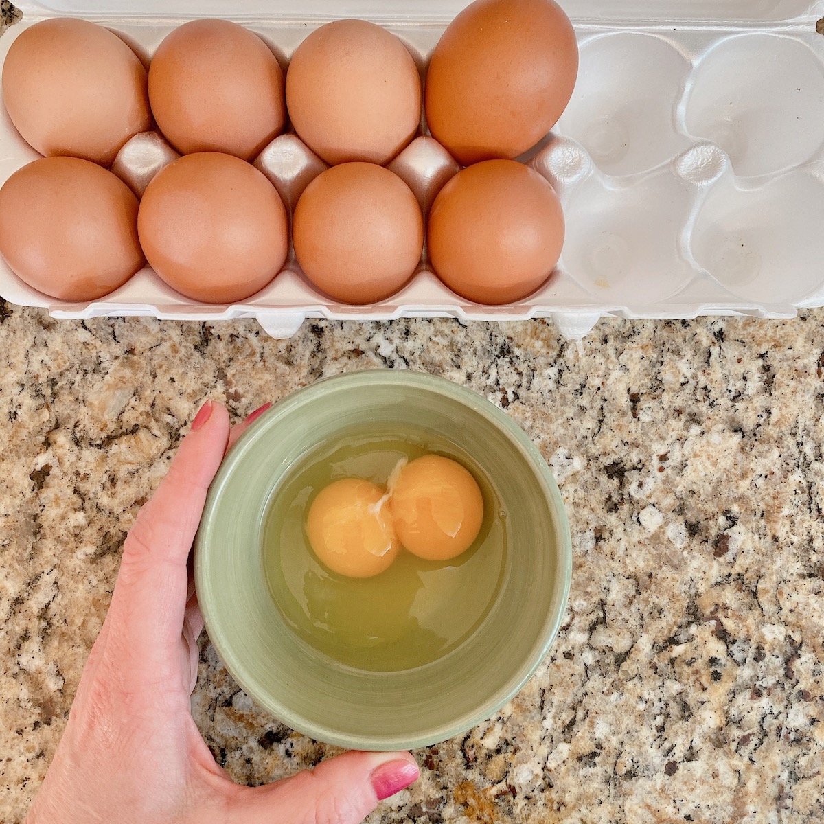 carton of eggs and a hand holding a bowl of a cracked double yolk egg